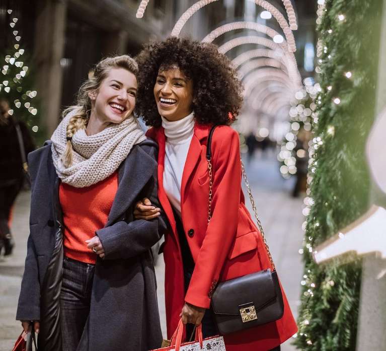 Two women wearing elegant jackets smile happily while holding gift bags in front of holiday lights
