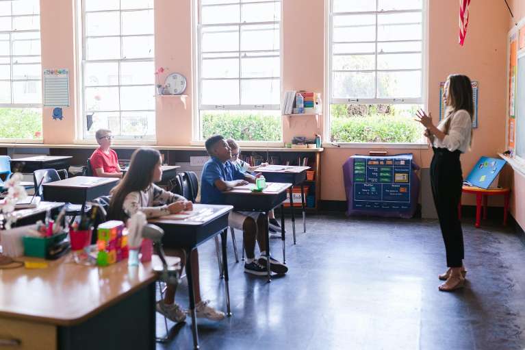 A teacher stands at the front of a classroom teaching young students sitting in desks